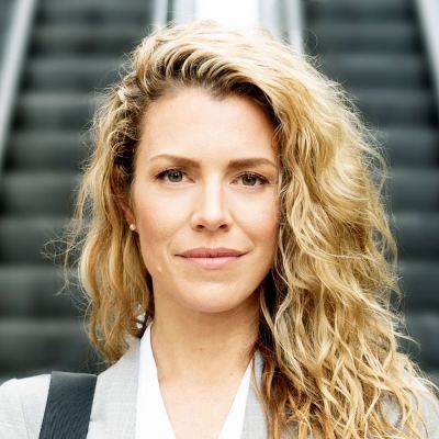 Smiling light-skinned, female-presenting individual with long blonde hair wearing a grey jacket in front of stairs